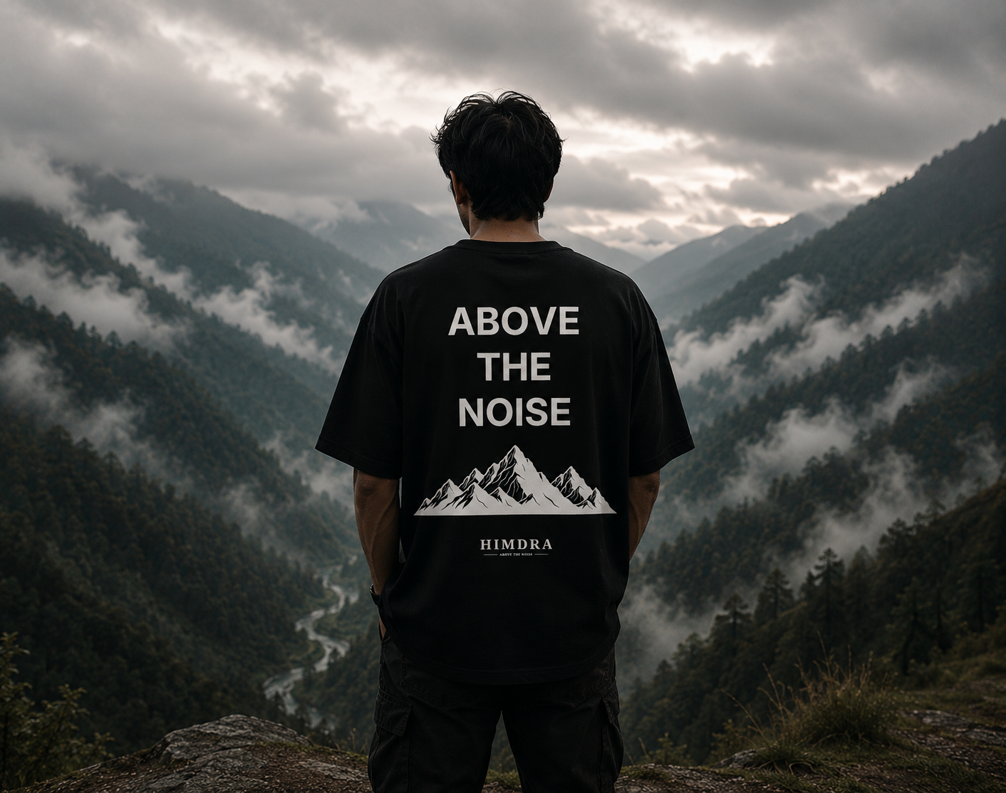 Person wearing a black t-shirt with 'Above the Noise' design, standing on a mountain with a scenic view.
