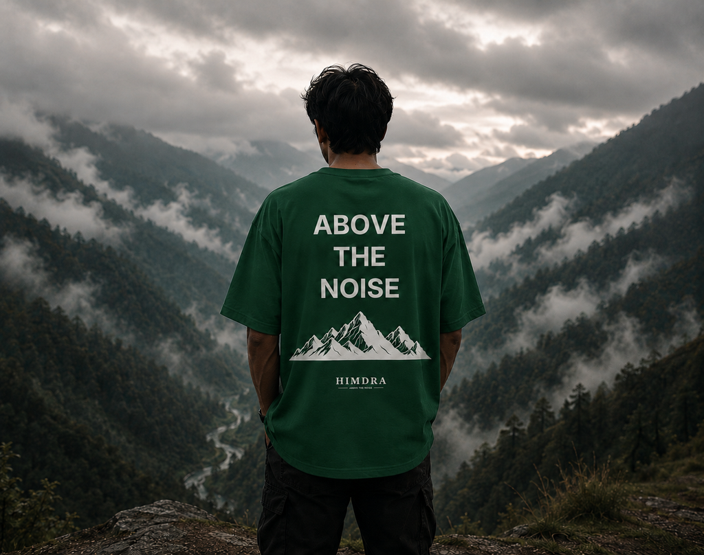 Person wearing a green t-shirt with 'Above the Noise' design, standing on a mountain with a scenic view.