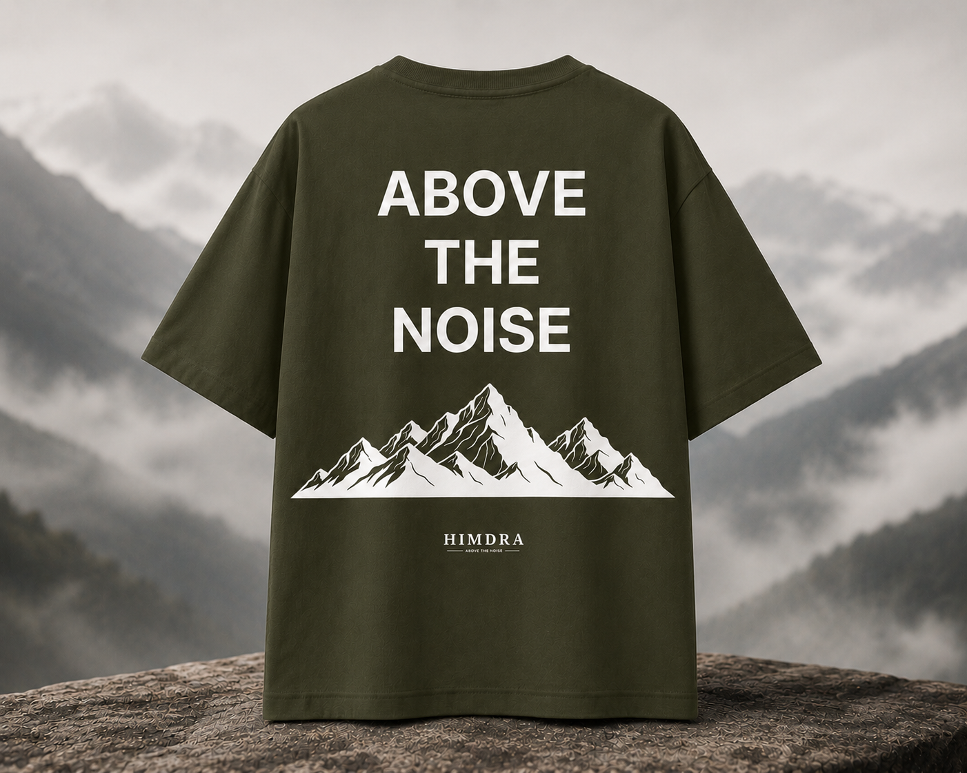Green t-shirt with 'Above the Noise' and mountain design on a rocky surface with mountain background
